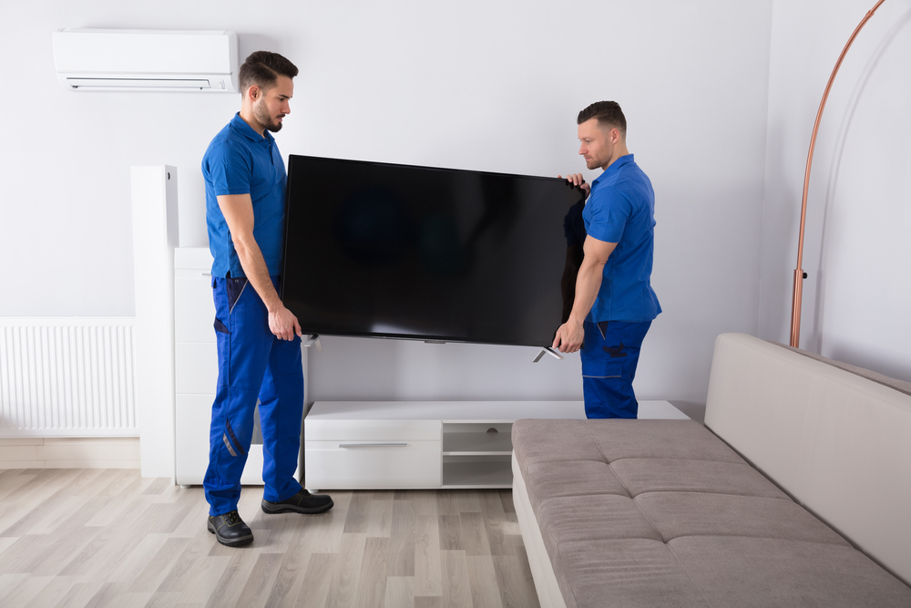 Two Young Male Movers In Uniform Placing Television In Living Room