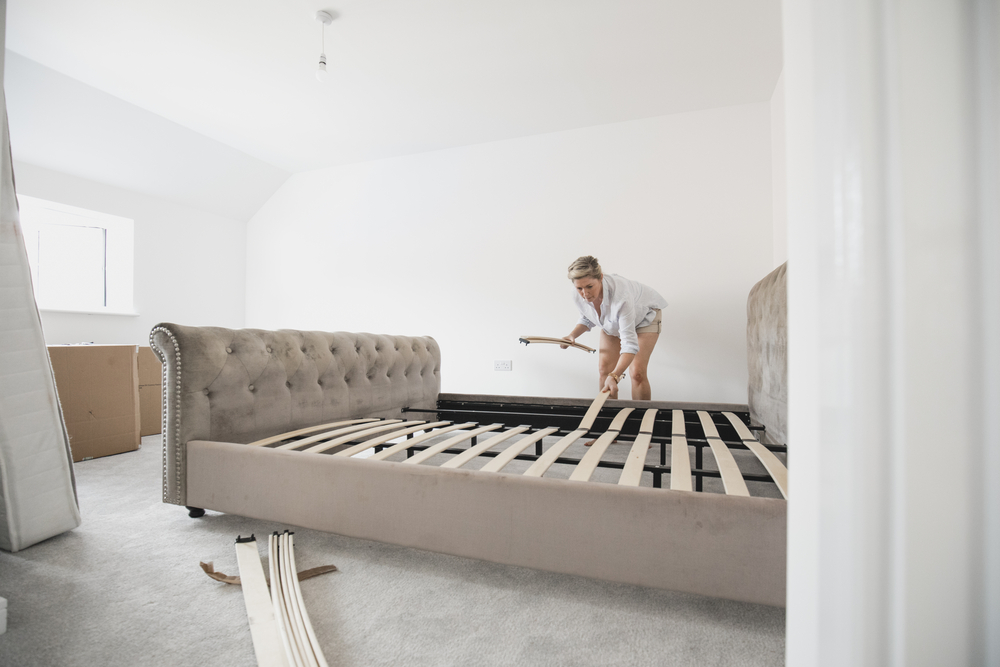 Mature woman is building her bed in her new home. She is placing wooden slats on the bed frame.