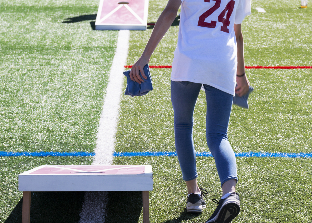 Rear view of a high school girl throwing bean bags while playing corn hole on a green turf field on a sunny day.