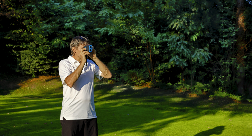 The golfer accurately measures the distance to the flag in a golf course with the laser rangefinder. In the background the forest.