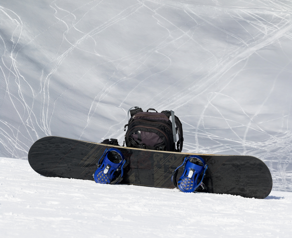 Snowboard and black backpack on snow in high winter mountains and snowy off-piste slope with traces of skis and snowboards at background